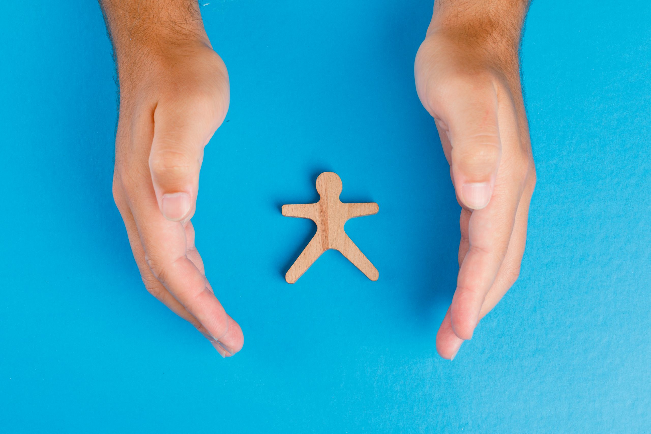 Social protection concept on blue background flat lay. hands taking care of wooden human figure.